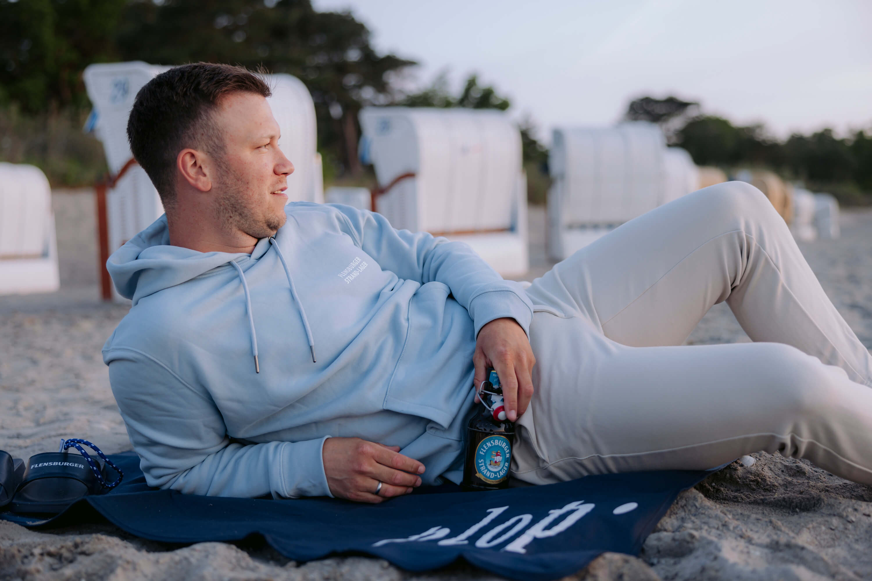 Ein Mann liegt auf einer navyblauen Stranddecke „Hvide Sande“ im Sand. Er trägt einen hellblauen Hoodie mit der Aufschrift „Flensburger Strand-Lager“ und hält eine Flasche Flensburger Strand-Lager Bier.