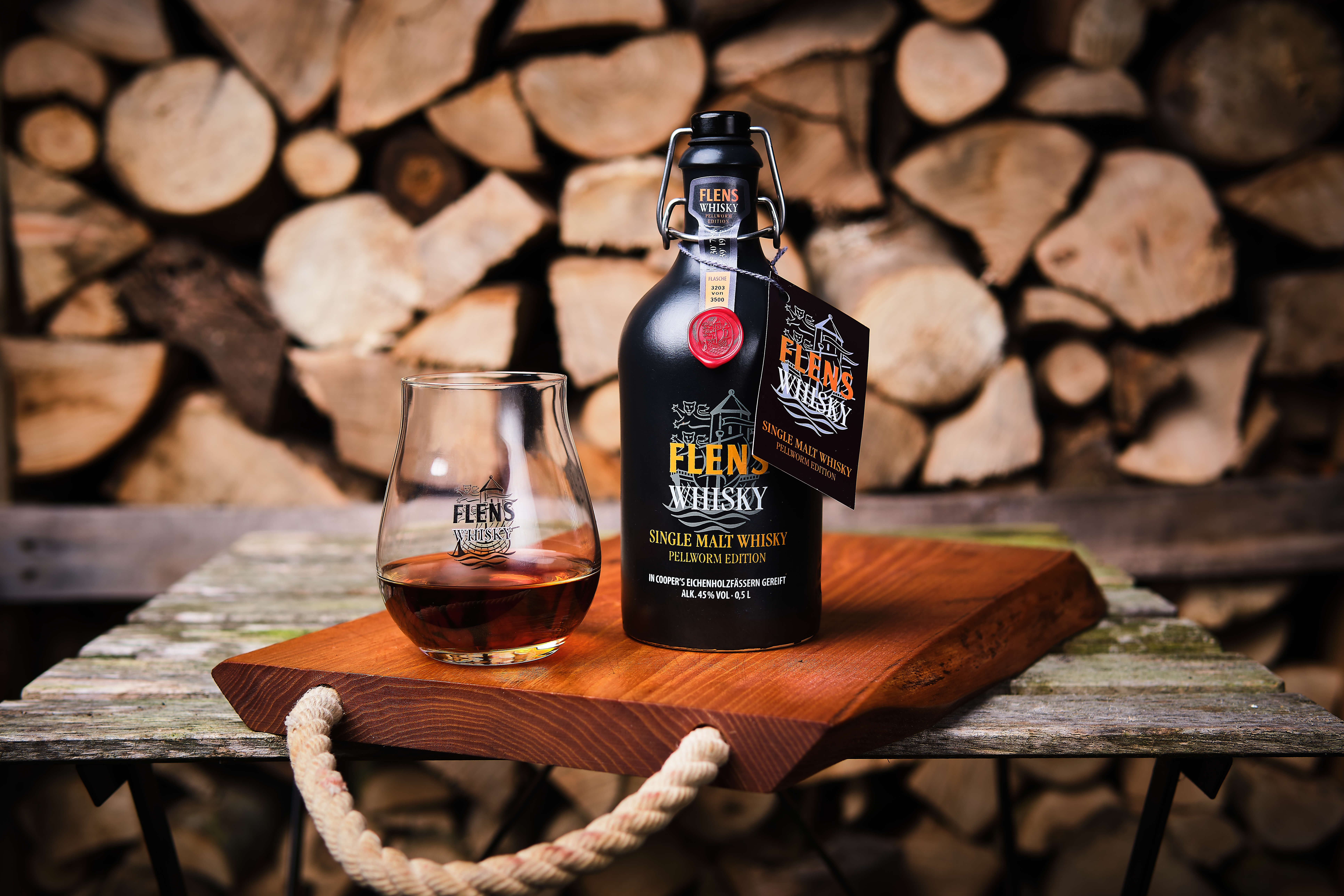 Flens Whisky Flasche "Single Malt Whisky Pellworm Edition" mit einem Glas teilgefüllt steht auf einem Holzbrett vor aufgeschichtetem Kaminholz.