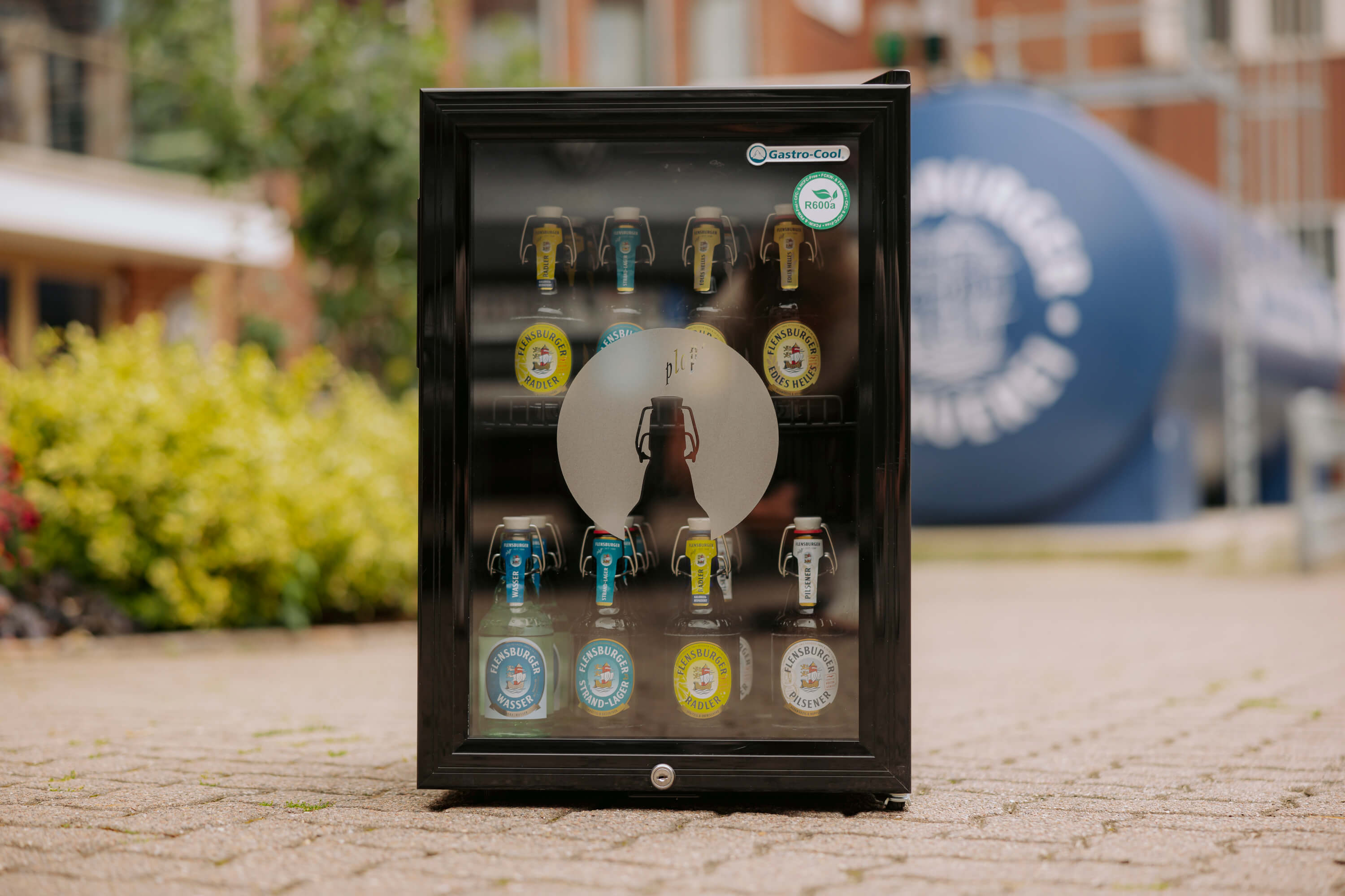 Schwarzer Tresenkühlschrank mit Glasfront zeigt mehrere Flaschen Flensburger Bier und Wasser mit Bügelverschlüssen. Auf der Tür steht ein großer, grauer Flaschen-Schatten und Aufkleber mit "Gastro-Cool".