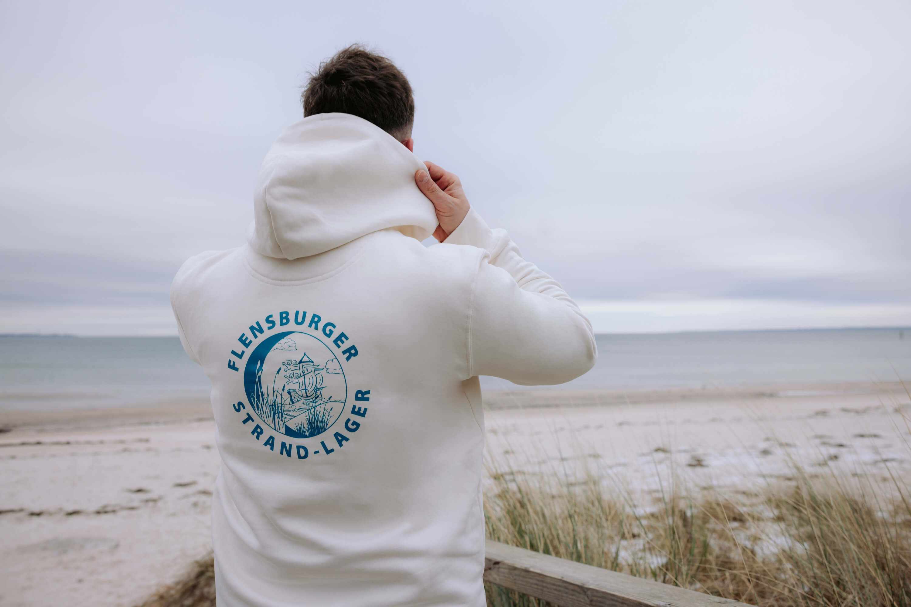 Person trägt beigen Hoodie mit blauem Logo und Schriftzug "Flensburger Strand-Lager" am Rücken und blickt auf einen Strand mit Gras und ruhigem Meer unter bewölktem Himmel.
