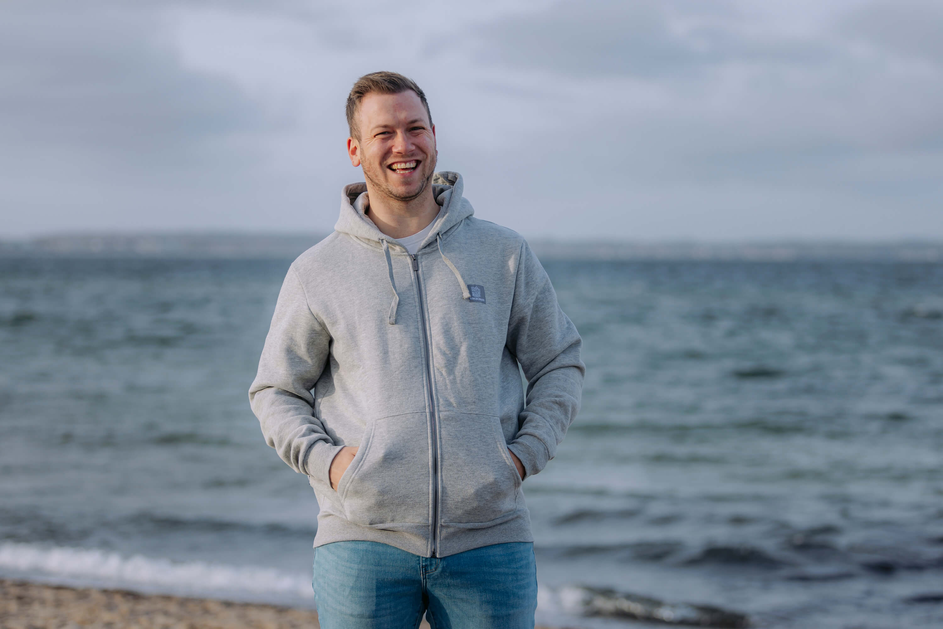 Ein Mann steht lächelnd am Strand. Er trägt eine hellgraue Sweatshirtjacke mit Reißverschluss und hellblaue Jeans. Im Hintergrund ist das Meer und der bewölkte Himmel zu sehen.