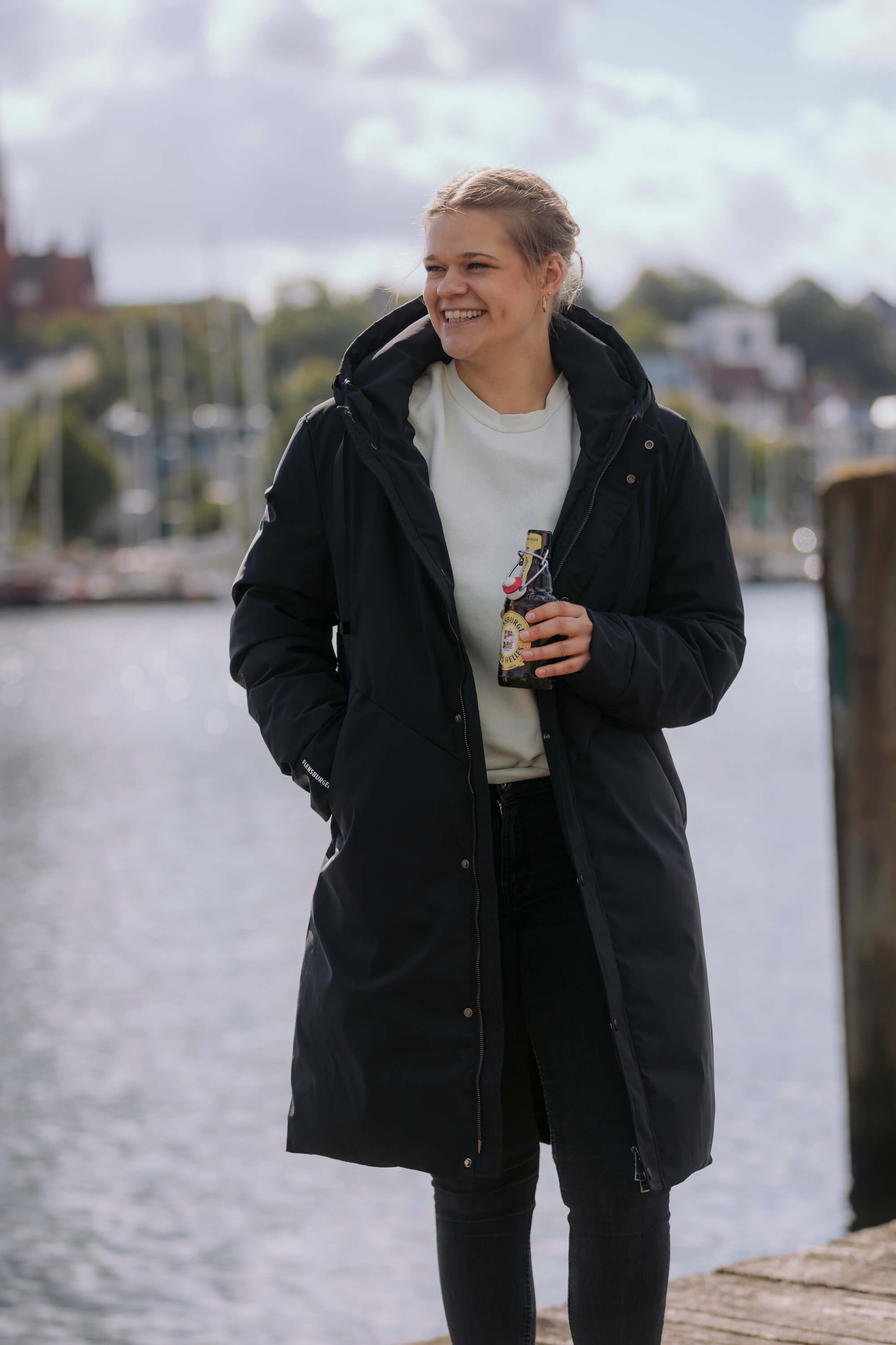 Frau trägt den FLENS Winterparka „Ruben“ in Schwarz. Sie hält eine Flasche Flensburger Pilsener und steht lachend am Wasser mit Booten im Hintergrund.